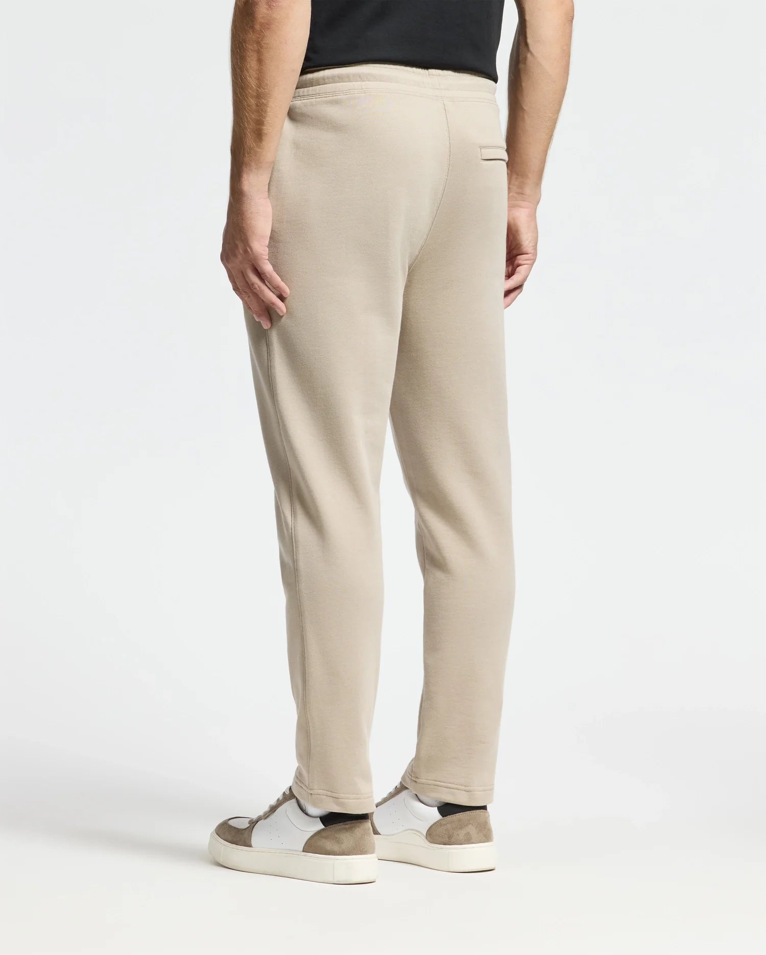A person is seen from behind wearing beige Monroe twill patch fleece sweatpants by Psycho Bunny and a black shirt. The look is complemented by white sneakers with brown accents against a plain white background, emphasizing the casual outfit.