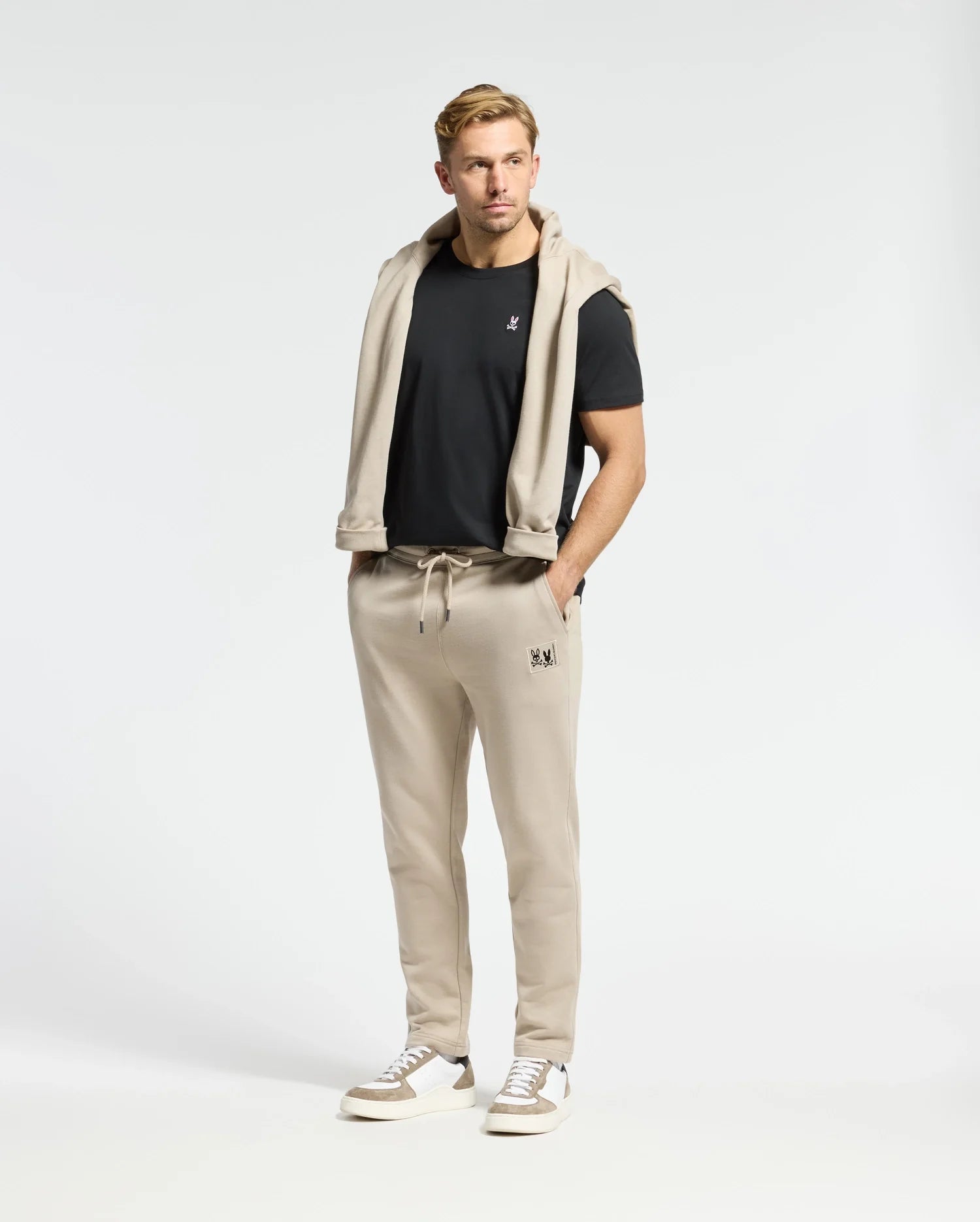 A man in a black T-shirt with a light beige sweater draped over his shoulders, wearing Psycho Bunny's Monroe Twill Patch Fleece Sweatpants and white sneakers, stands against a plain white background.