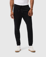 A person is wearing the MENS PACIFIC CHENILLE JOGGER - B6P972D200 by Psycho Bunny, featuring an adjustable drawstring and a laid-back bottom style, combined with white sneakers. Their ensemble is complemented by a chenille logo patch that adds subtle detail as their hands rest calmly at their sides, set against a light gray background.