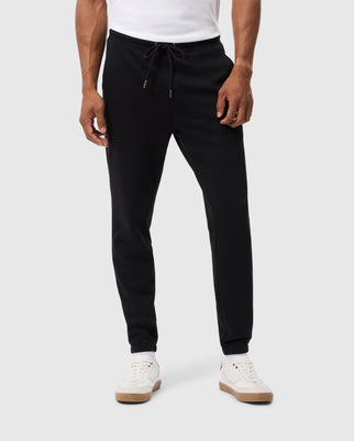 A person is wearing the MENS PACIFIC CHENILLE JOGGER - B6P972D200 by Psycho Bunny, featuring an adjustable drawstring and a laid-back bottom style, combined with white sneakers. Their ensemble is complemented by a chenille logo patch that adds subtle detail as their hands rest calmly at their sides, set against a light gray background.