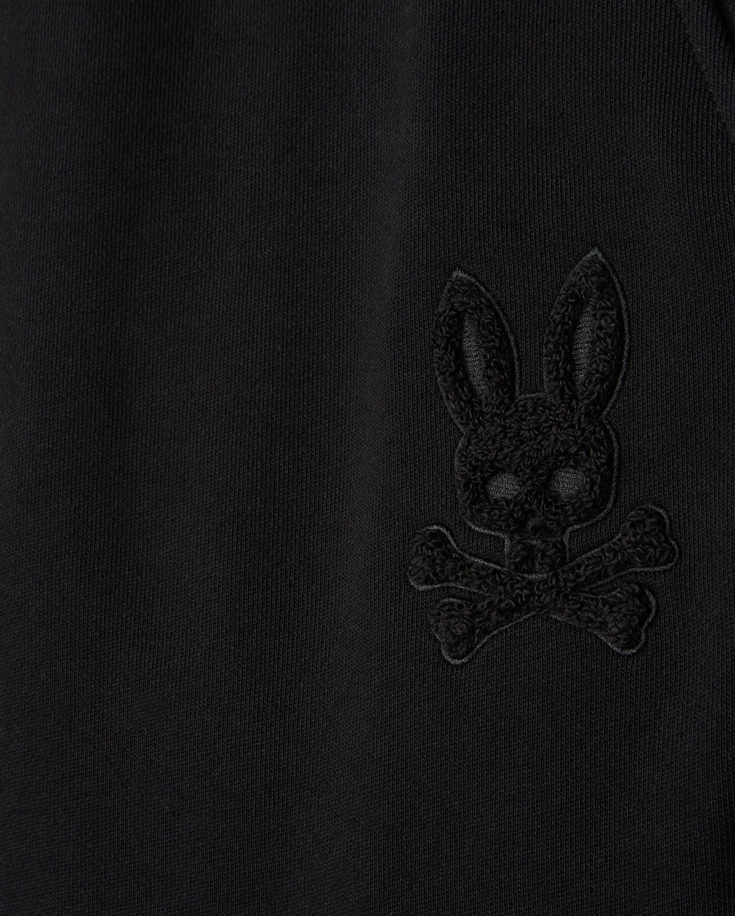 A close-up of the black fabric on our Psycho Bunny MENS PACIFIC CHENILLE JOGGER - B6P972D200 showcases an embroidered design featuring a skull and crossbones with a rabbit face and long ears, perfectly complementing the laid-back vibe of our casual jogger line.