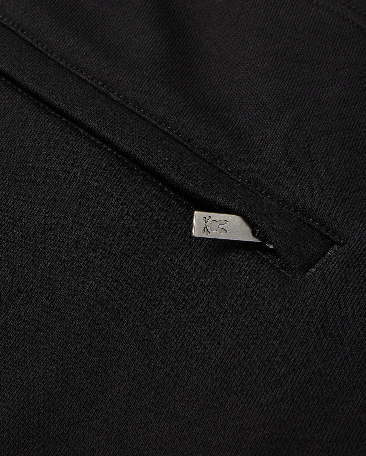 Close-up of a black fabric showcasing a small silver zipper pull with an engraved rabbit design. The zipper is partially open, providing a glimpse of the Psycho Bunny MENS PACIFIC CHENILLE JOGGER - B6P972D200 underneath, highlighting its casual bottom and unique chenille logo patch.