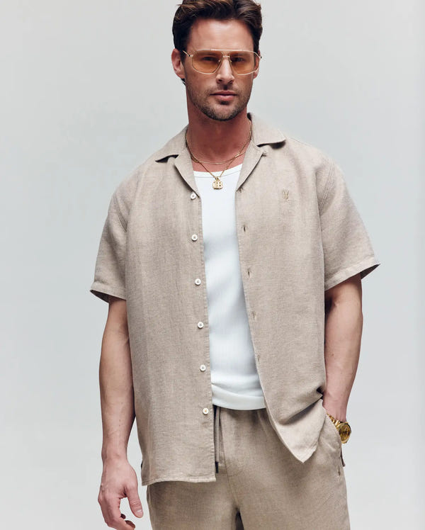 A man stands with one hand in his pocket, wearing Psycho Bunny’s MENS WINDCREST LINEN BLEND SHORT SLEEVE SHIRT (B6Q148C200) in beige over a white ribbed tank, paired with sunglasses, gold necklaces, a watch, and beige pants against a plain background.