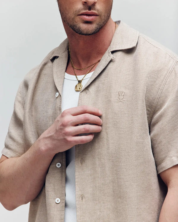 A man wears the Psycho Bunny MENS WINDCREST LINEN BLEND SHORT SLEEVE SHIRT (B6Q148C200) in beige over a white ribbed tee, accessorized with gold necklaces. His face is partly visible as he touches his shirt near the chest.