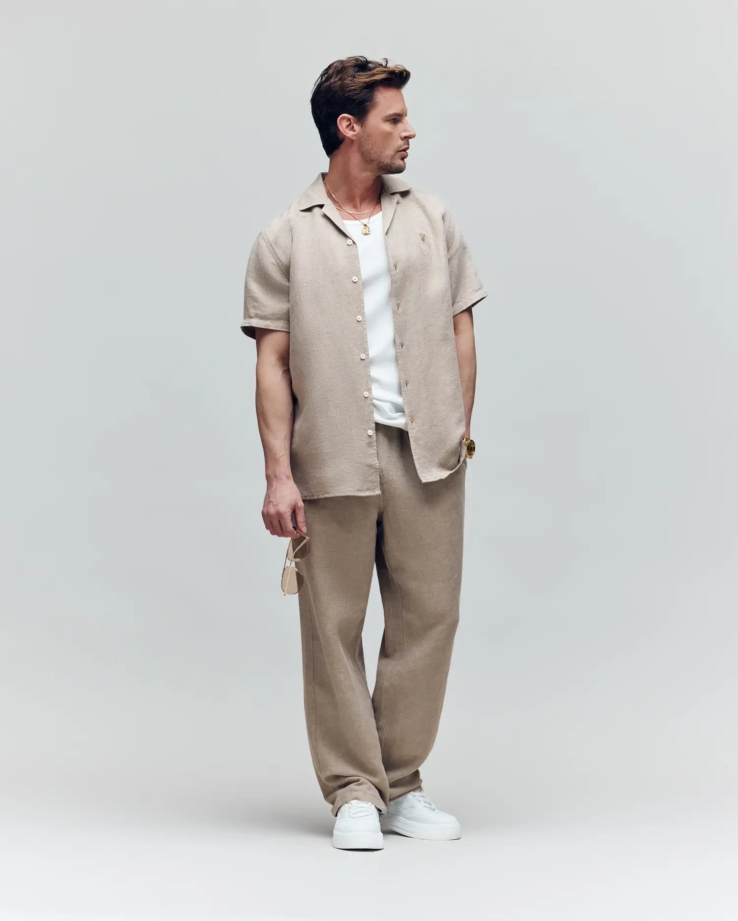 A man wears the Psycho Bunny MENS WINDCREST LINEN BLEND SHORT SLEEVE SHIRT (B6Q148C200) in beige over a white tee, with matching beige pants and white sneakers. He holds sunglasses and looks left, set against a plain light background.