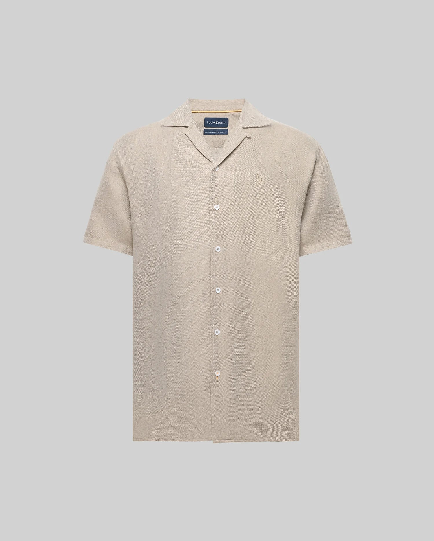 The Psycho Bunny MENS WINDCREST LINEN BLEND SHORT SLEEVE SHIRT (B6Q148C200) is a beige, linen-cotton button-up with short sleeves, notched collar, chest pocket, embroidered logo on the left, and mother-of-pearl buttons against a light gray background.
