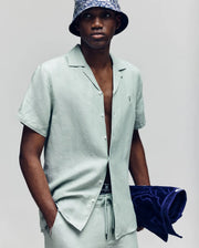 A man wearing the Psycho Bunny MENS WINDCREST LINEN BLEND SHORT SLEEVE SHIRT (B6Q148C200) in light green with matching pants stands facing forward, holding a navy towel and finishing his look with a dark patterned bucket hat.