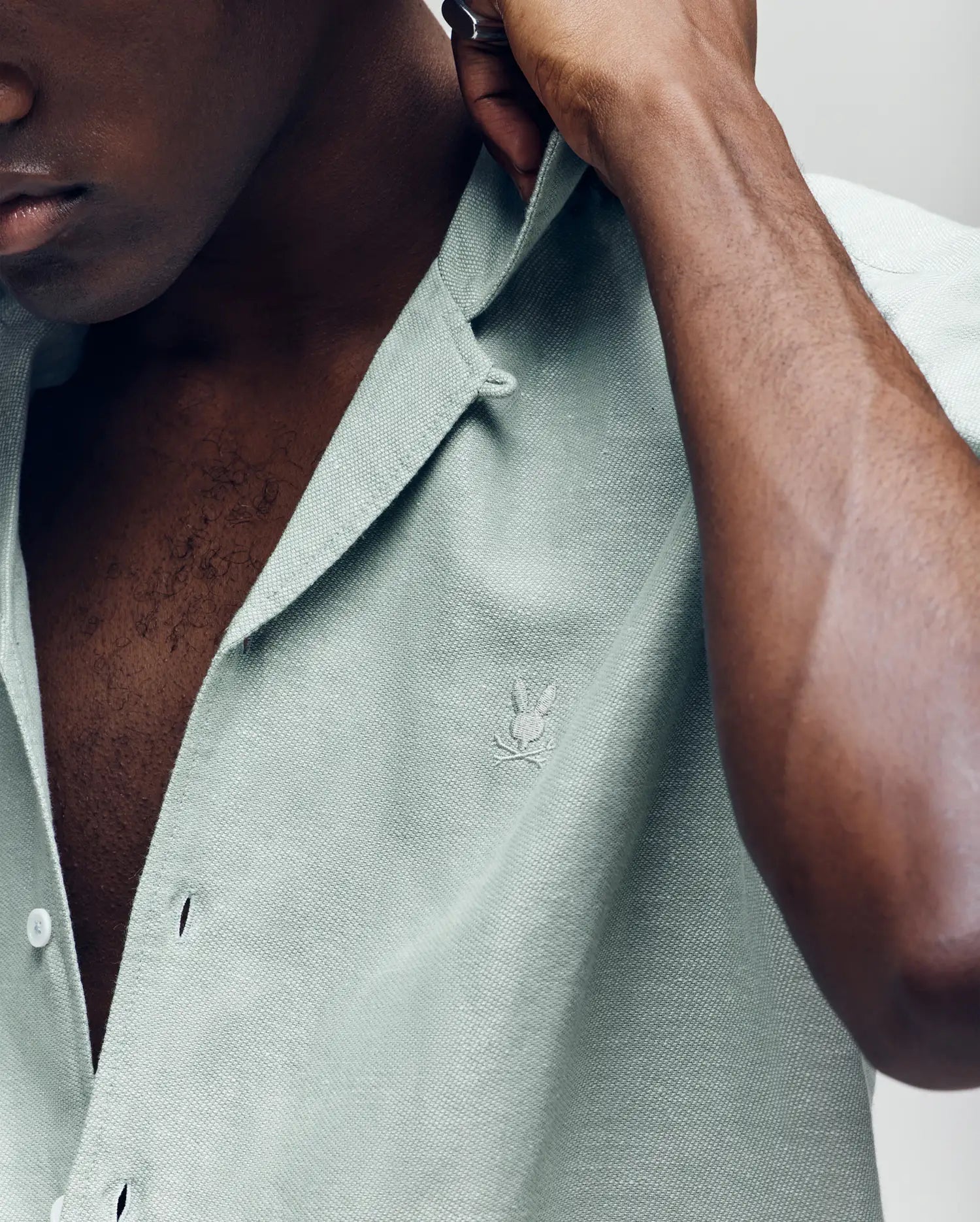 A close-up of a person buttoning the Psycho Bunny MENS WINDCREST LINEN BLEND SHORT SLEEVE SHIRT, featuring a light green linen-cotton blend, open spread collar, chest logo embroidery, and Mother-of-Pearl buttons.