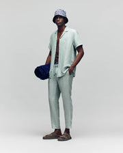 A man in a Psycho Bunny MENS WINDCREST LINEN BLEND SHORT SLEEVE SHIRT (B6Q148C200) in light sage green, with matching drawstring pants, bucket hat, sandals, and holding a navy beach towel stands against a plain gray background.