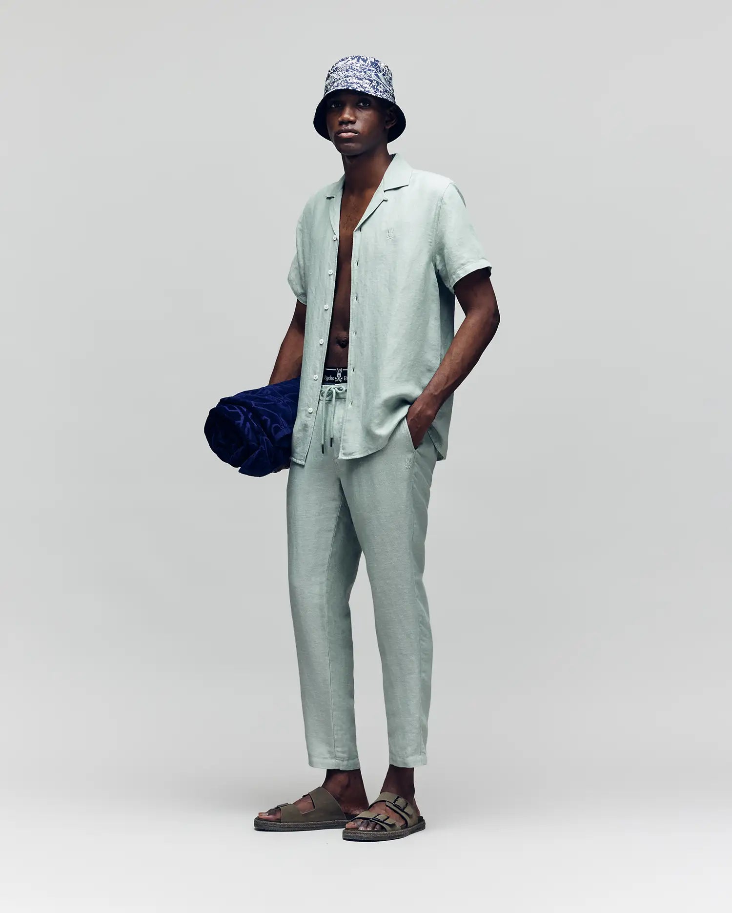 A man in a Psycho Bunny MENS WINDCREST LINEN BLEND SHORT SLEEVE SHIRT (B6Q148C200) in light sage green, with matching drawstring pants, bucket hat, sandals, and holding a navy beach towel stands against a plain gray background.