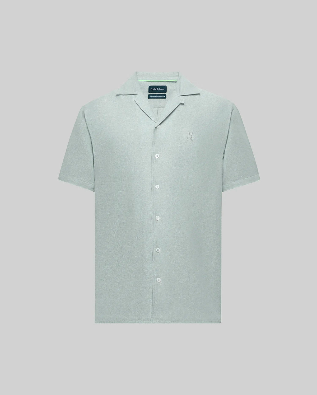 The Psycho Bunny MENS WINDCREST LINEN BLEND SHORT SLEEVE SHIRT (B6Q148C200) is a light green button-up with short sleeves, open spread collar, white buttons, and a small embroidered logo on the left chest, set against a light gray background.