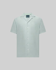 The Psycho Bunny MENS WINDCREST LINEN BLEND SHORT SLEEVE SHIRT (B6Q148C200) is a light green button-up with short sleeves, open spread collar, white buttons, and a small embroidered logo on the left chest, set against a light gray background.