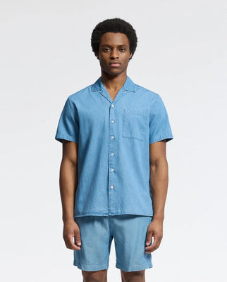 A person stands facing forward, wearing the Psycho Bunny MENS KERVIN LIGHTWEIGHT DENIM WOVEN SHIRT - B6Q467F200, a casual short-sleeve denim shirt with a pocket and matching shorts. A plain white background highlights the ensemble's stylish simplicity.