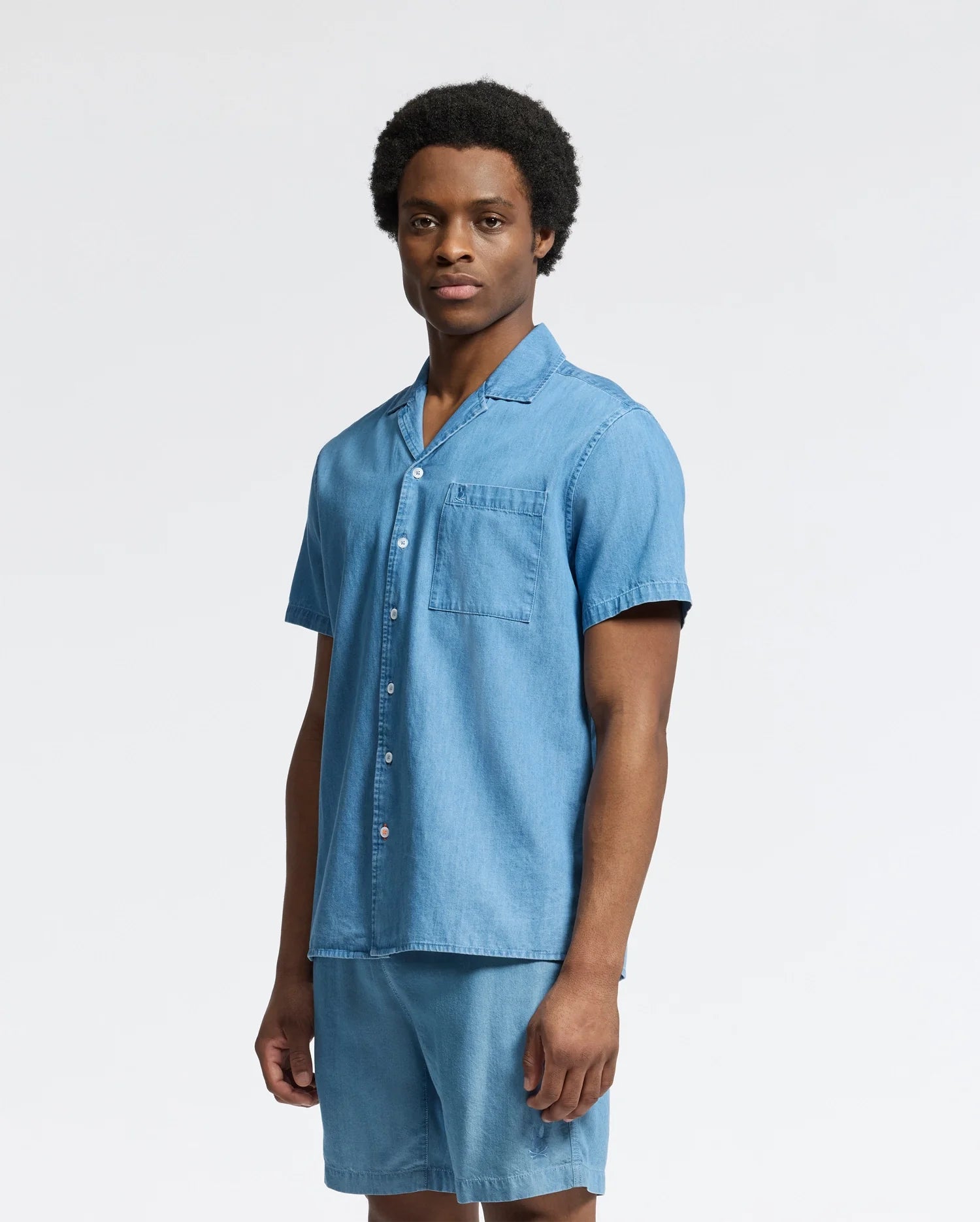 A person confidently models the Psycho Bunny MENS KERVIN LIGHTWEIGHT DENIM WOVEN SHIRT (B6Q467F200), paired with matching shorts, against a plain white background.