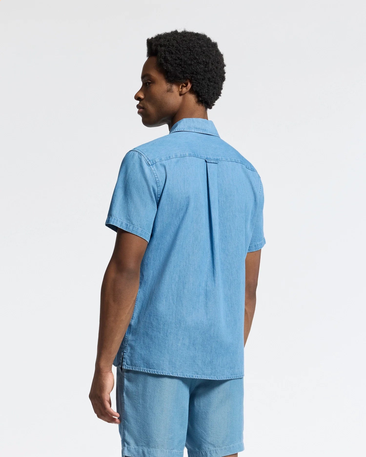 A person stands against a plain light background wearing the Psycho Bunny MENS KERVIN LIGHTWEIGHT DENIM WOVEN SHIRT in blue, with short sleeves and matching shorts, looking to the side.