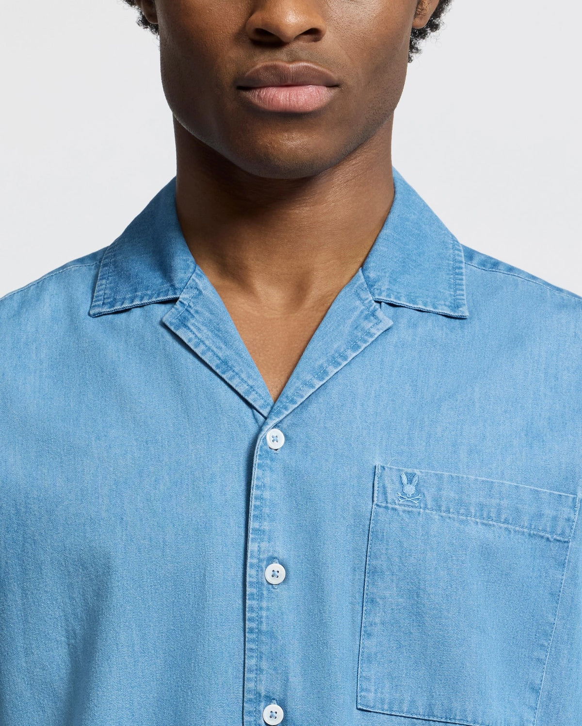 A man wears the Psycho Bunny MENS KERVIN LIGHTWEIGHT DENIM WOVEN SHIRT in light blue, with a collar and chest pocket. The image highlights his upper body against a plain white background.