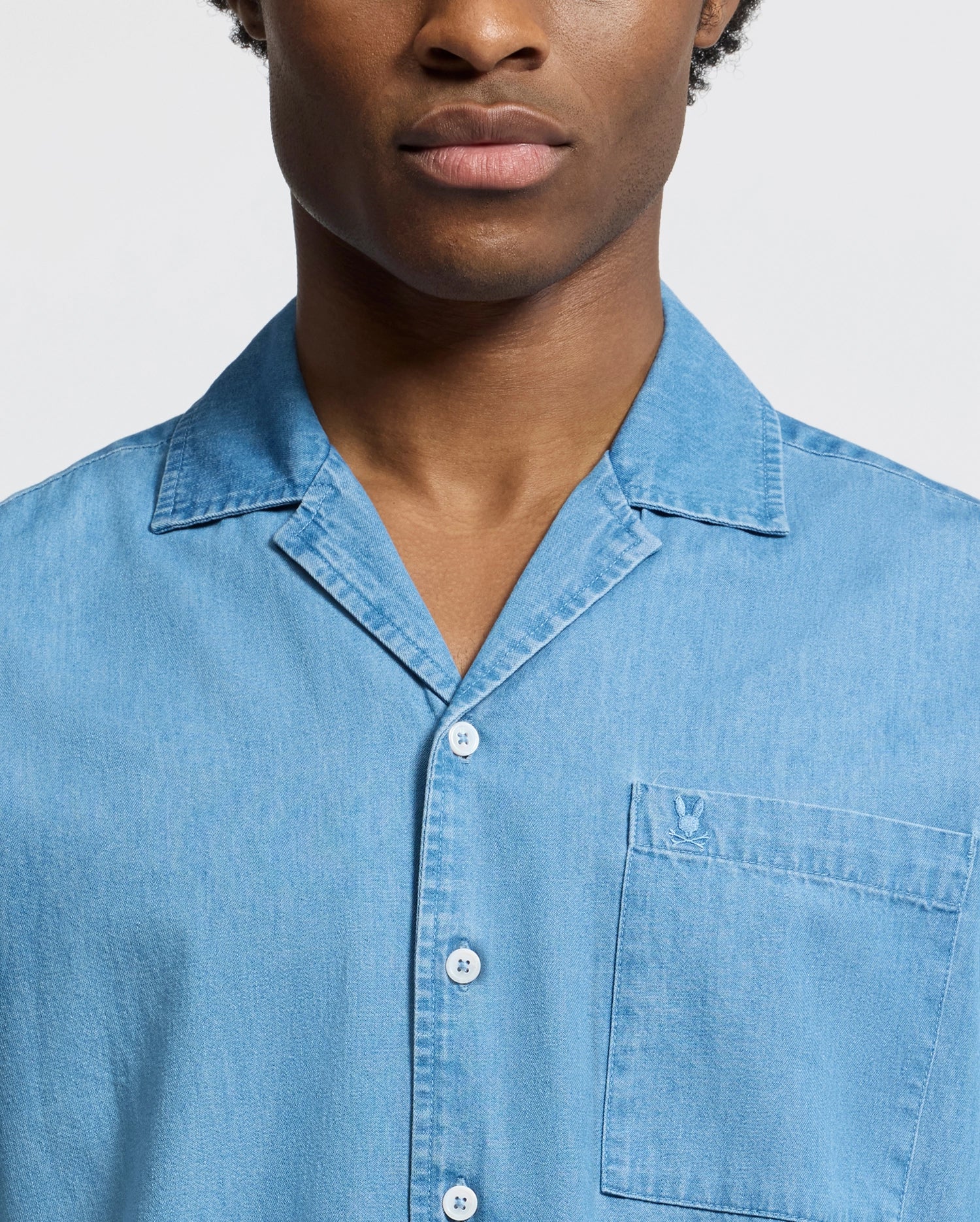 A man wears the Psycho Bunny MENS KERVIN LIGHTWEIGHT DENIM WOVEN SHIRT in light blue, with a collar and chest pocket. The image highlights his upper body against a plain white background.