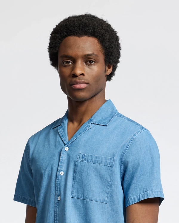 A person with short curly hair wears the MENS KERVIN LIGHTWEIGHT DENIM WOVEN SHIRT by Psycho Bunny, exuding an understated style. They stand against a plain, light background with a neutral expression.