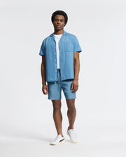 A man in a Psycho Bunny MENS KERVIN LIGHTWEIGHT DENIM WOVEN SHIRT, paired with matching shorts, exudes casual vibes on a white backdrop. A white tee and sneakers add understated elegance. His neutral expression and casually resting hands complete the look.