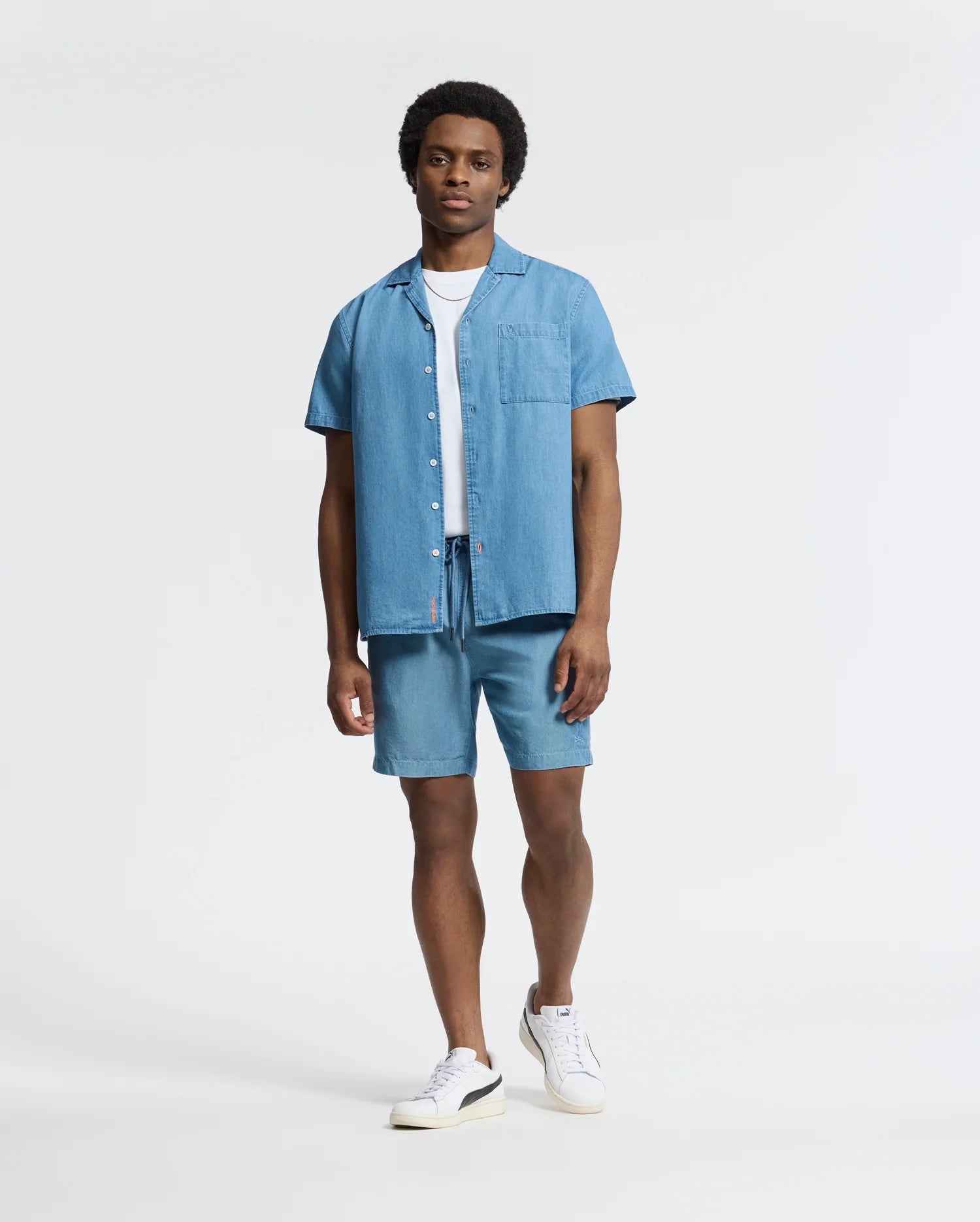 A man in a Psycho Bunny MENS KERVIN LIGHTWEIGHT DENIM WOVEN SHIRT, paired with matching shorts, exudes casual vibes on a white backdrop. A white tee and sneakers add understated elegance. His neutral expression and casually resting hands complete the look.