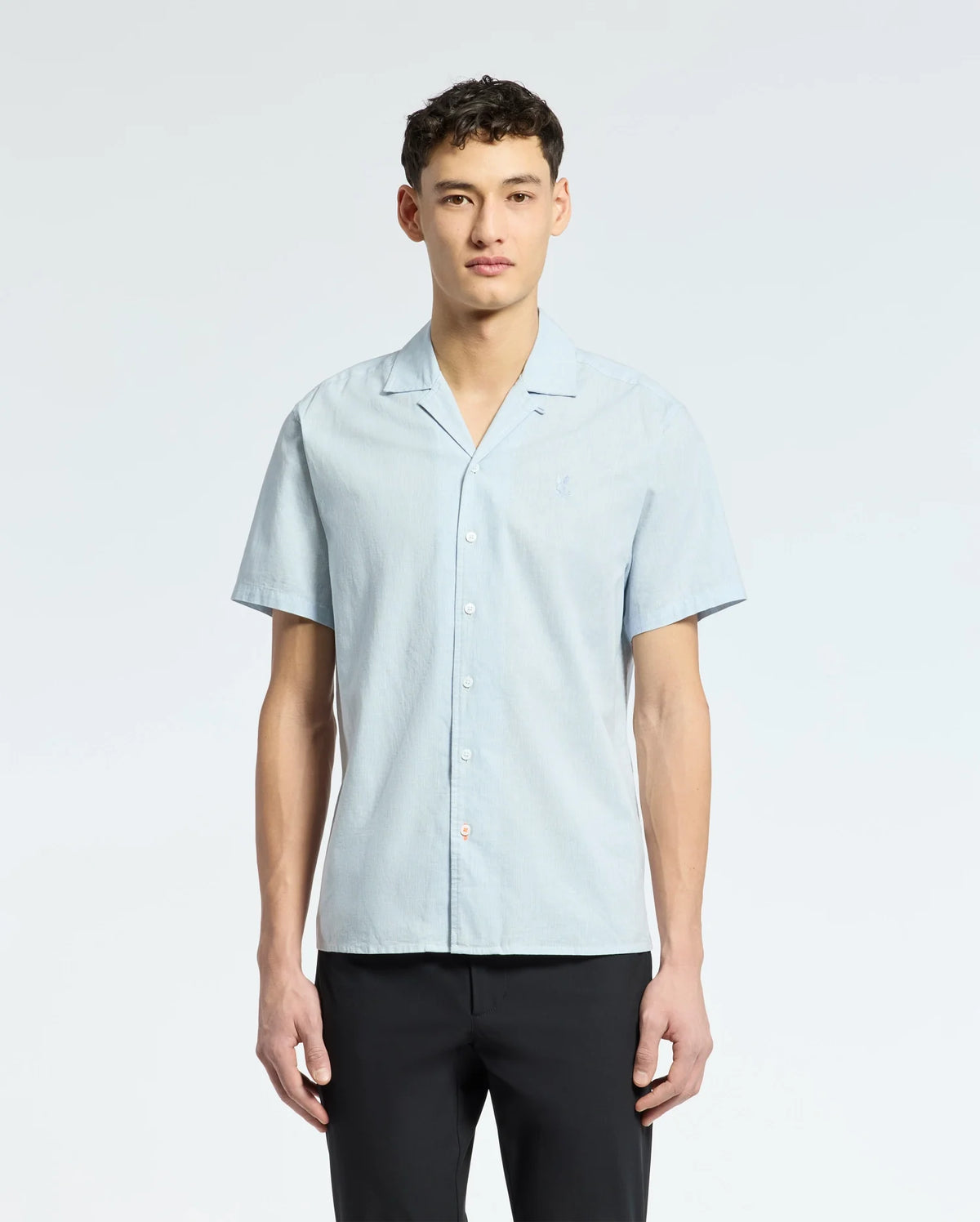 A young man stands facing forward against a plain white background, wearing the Psycho Bunny MENS COBAR WOVEN SHIRT - B6Q522F200 in light blue with tonal embroidery, paired with black pants.