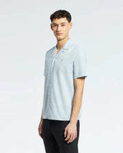 A man stands against a plain, light background wearing the Psycho Bunny MENS COBAR WOVEN SHIRT - B6Q522F200 in light blue with black pants, looking calmly at the camera with a neutral expression.