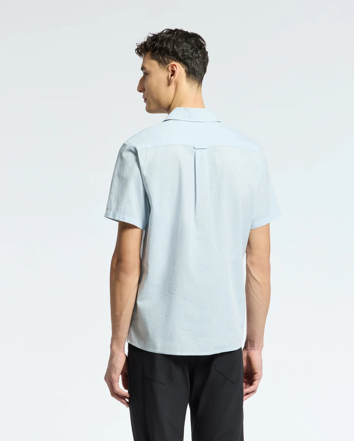 A man stands with his back to the camera in black pants and the Psycho Bunny MENS COBAR WOVEN SHIRT - B6Q522F200, against a plain light background.