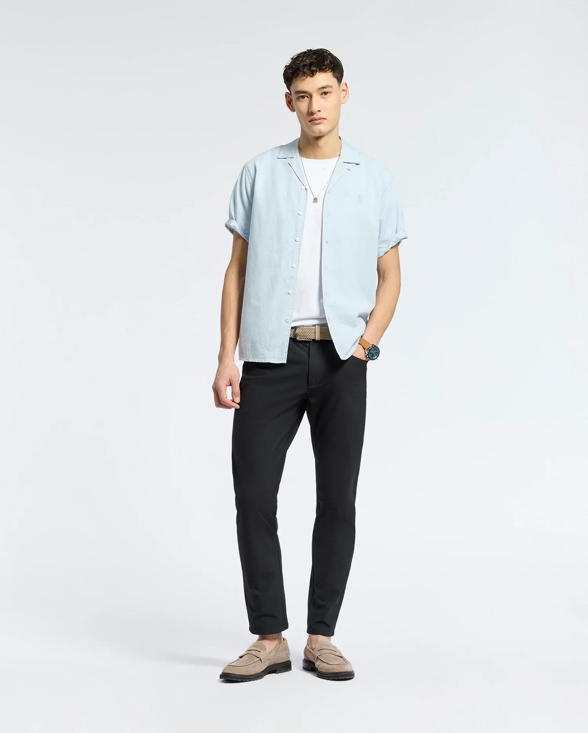 A man poses against a plain background wearing the Psycho Bunny MENS COBAR WOVEN SHIRT - B6Q522F200 over a white tee, with black pants, beige loafers, a belt, and a wristwatch. He faces the camera in a relaxed stance.