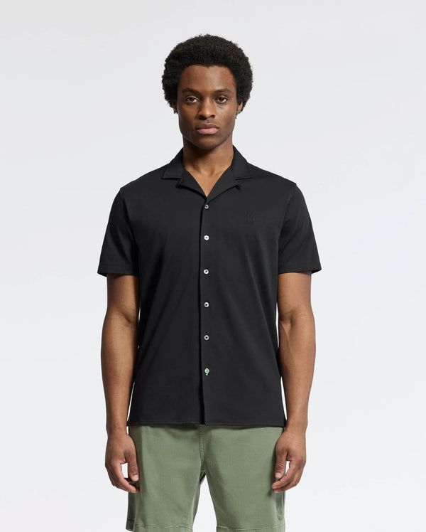 The person wears Psycho Bunny's MENS BRYAN CAMP COLLAR SHORT SLEEVE SHIRT - B6Q581F200, a black Pima cotton blend with a small logo on the left chest, and green shorts against a plain white background. They have short curly hair and a neutral expression.