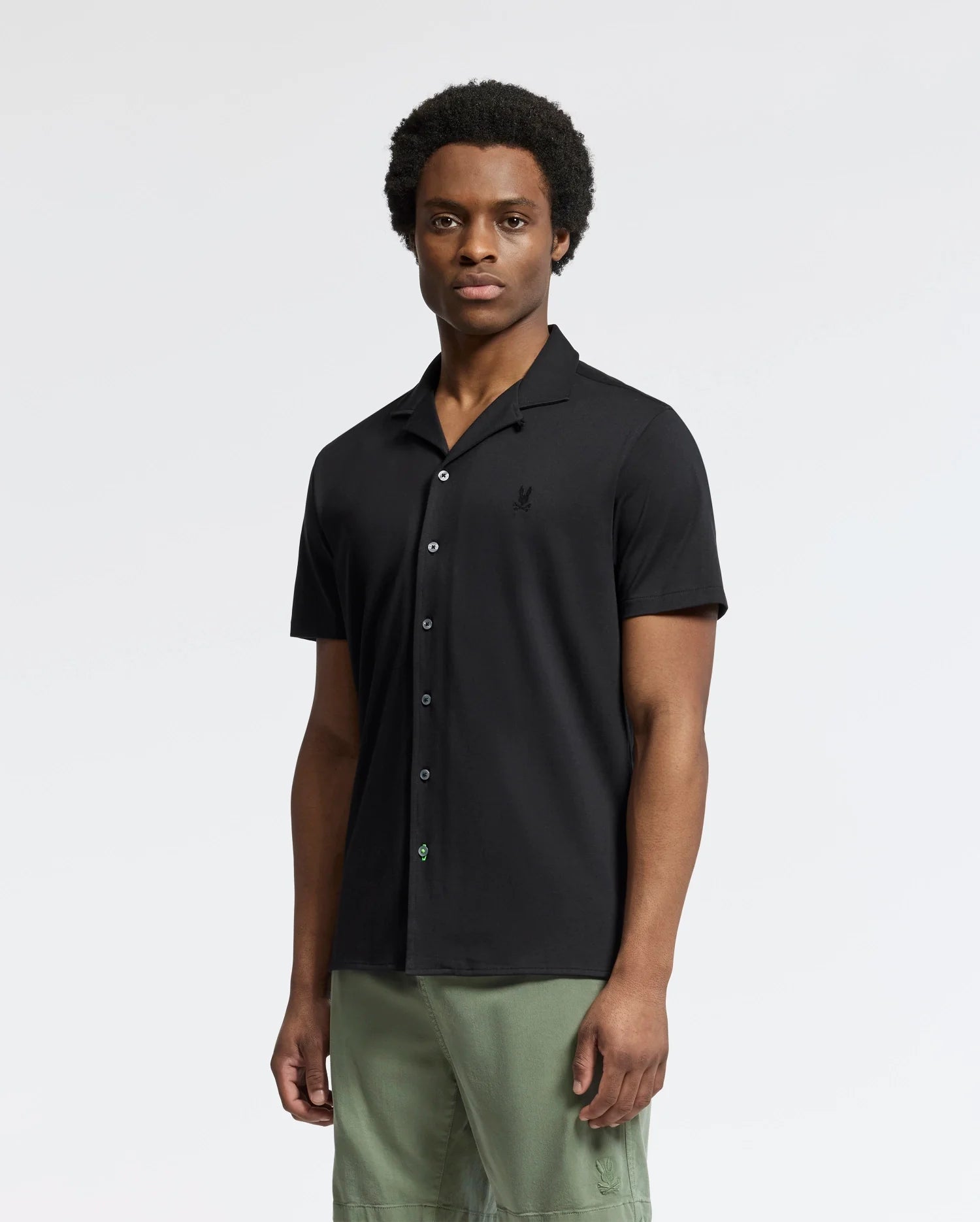 A person with short curly hair wears a Psycho Bunny MENS BRYAN CAMP COLLAR SHORT SLEEVE SHIRT in black and green pants while standing against a plain white background, facing the camera with a neutral expression.