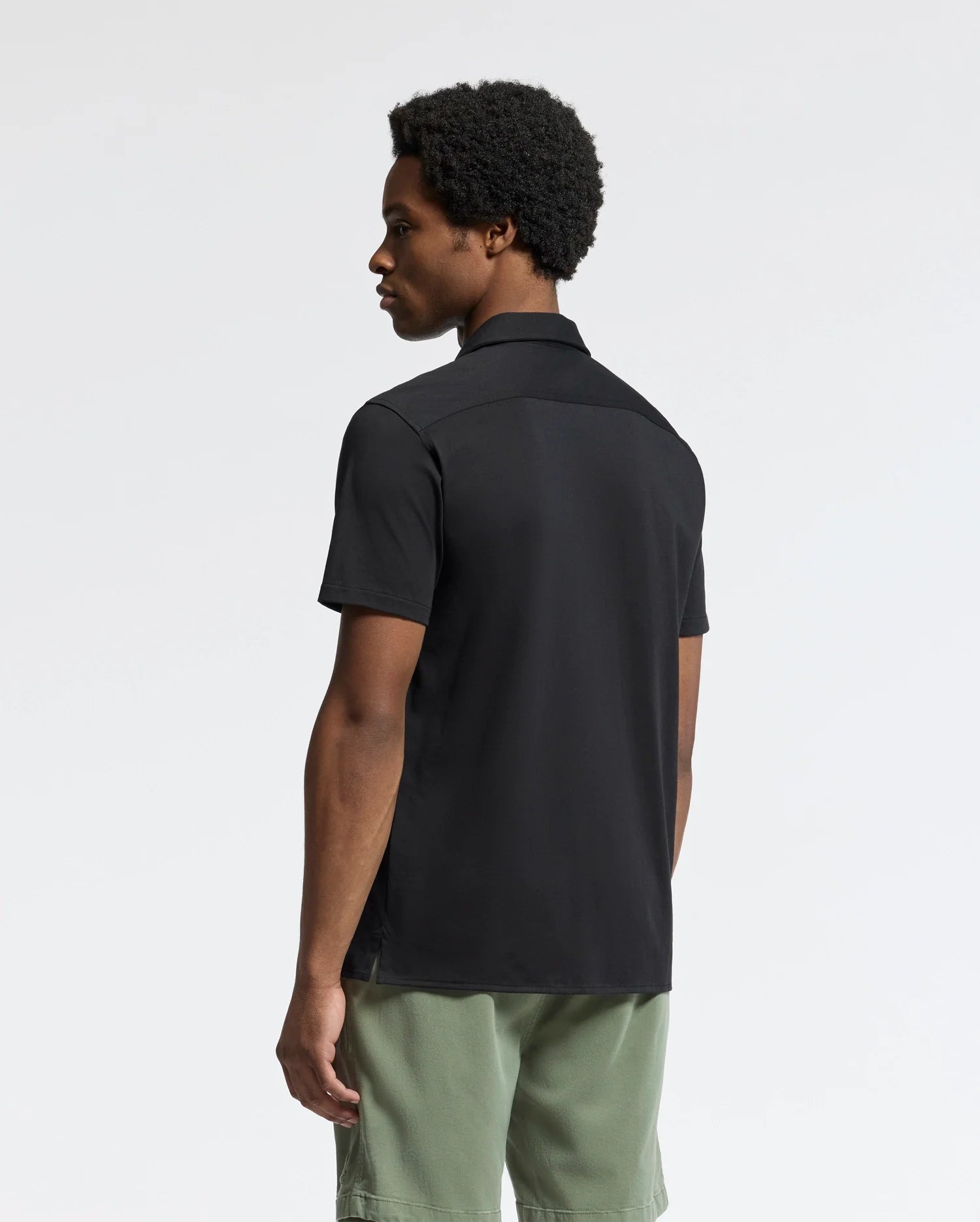 A person with short curly hair stands sideways, wearing a Psycho Bunny mens' Bryan Camp Collar Short Sleeve Shirt in black, made of moisture-wicking Pima cotton blend, paired with light green shorts. The background is plain and light gray.