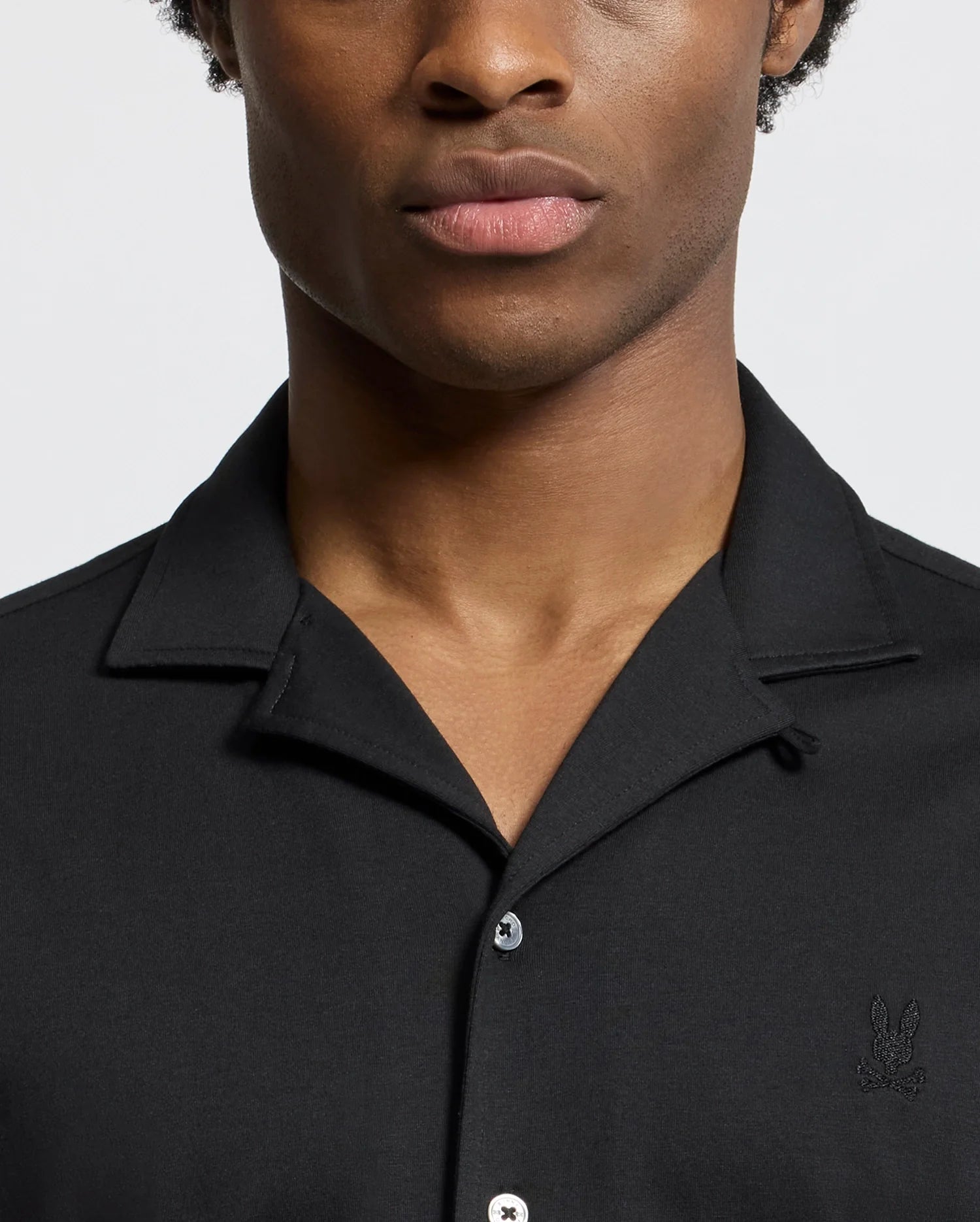 A person wearing the Psycho Bunny MENS BRYAN CAMP COLLAR SHORT SLEEVE SHIRT - B6Q581F200, featuring a black collared design with an embroidered emblem and crafted from a Pima cotton blend for ultimate comfort, is facing forward against a plain background, cropped from shoulders to chin.