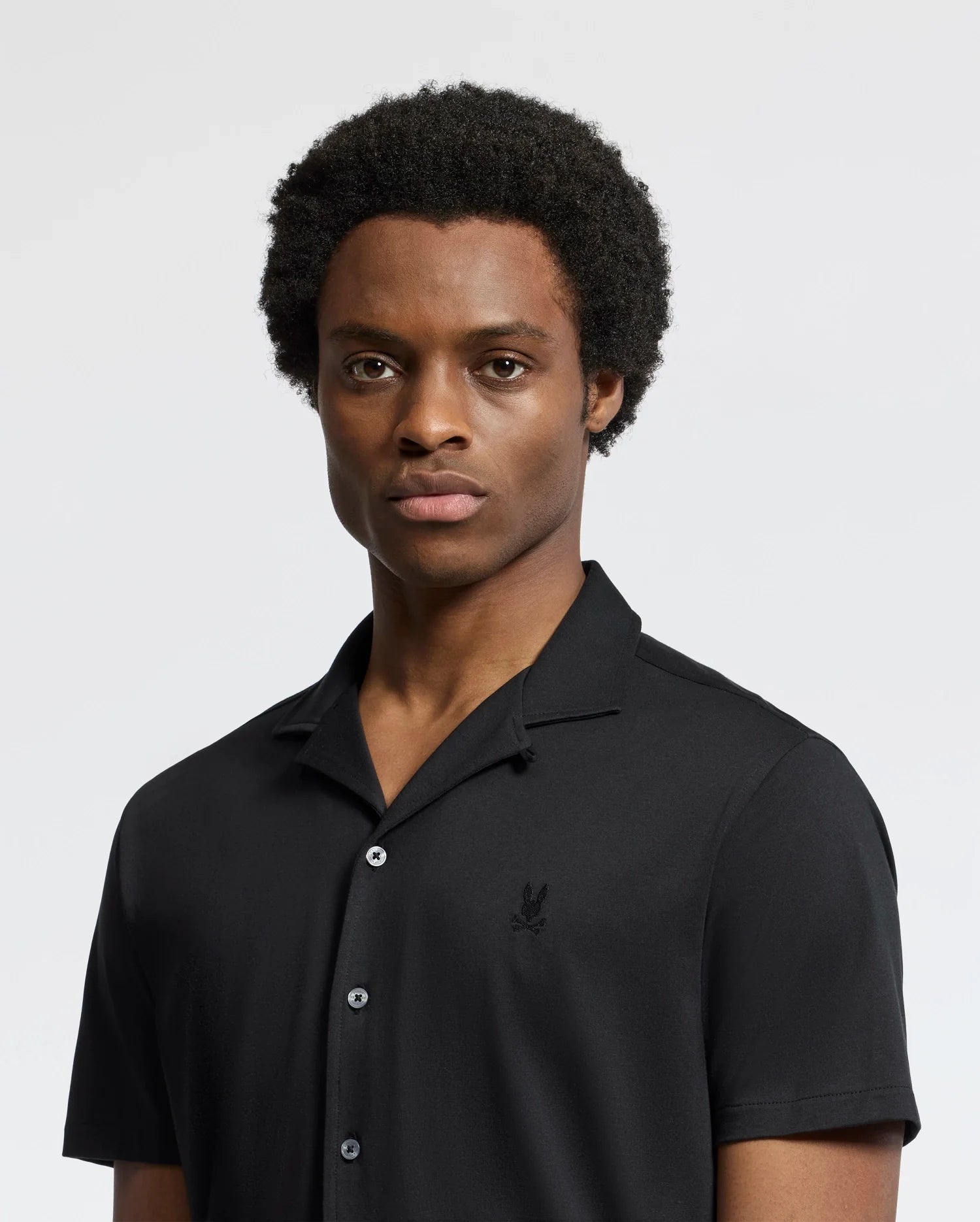 A person with short curly hair is wearing the Psycho Bunny MENS BRYAN CAMP COLLAR SHORT SLEEVE SHIRT in black, made from a Pima cotton blend. They stand against a plain light background, looking directly at the camera with a neutral expression.