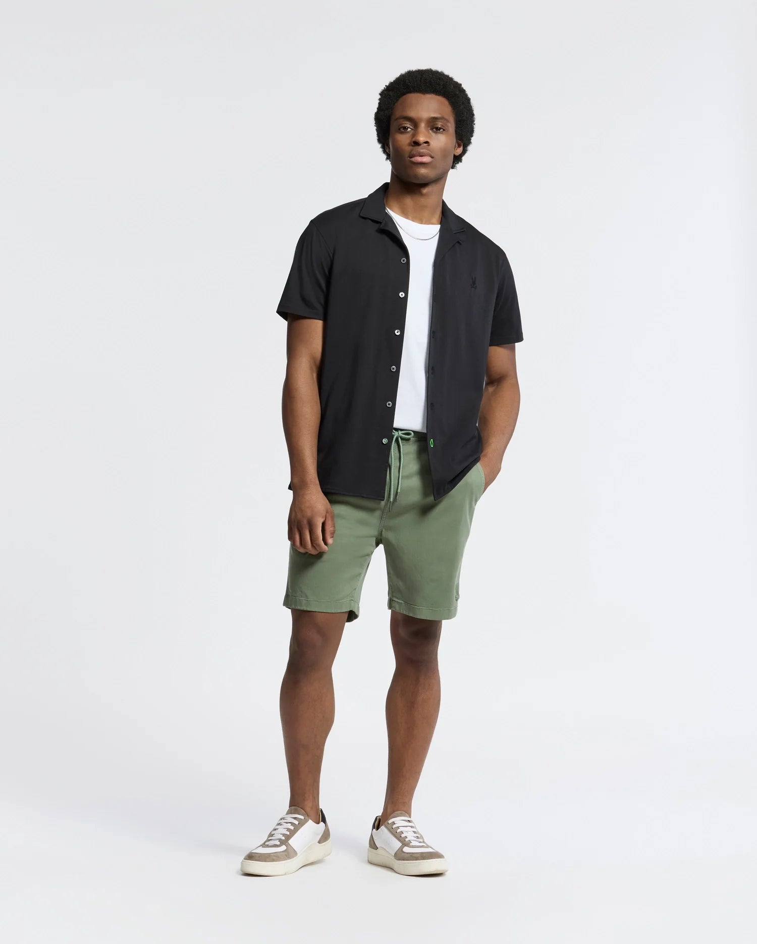 Confidently standing in a Psycho Bunny black short-sleeve camp collar shirt over a white T-shirt, paired with green shorts and beige sneakers, they casually pocket one hand against a plain white background, embodying effortless style.