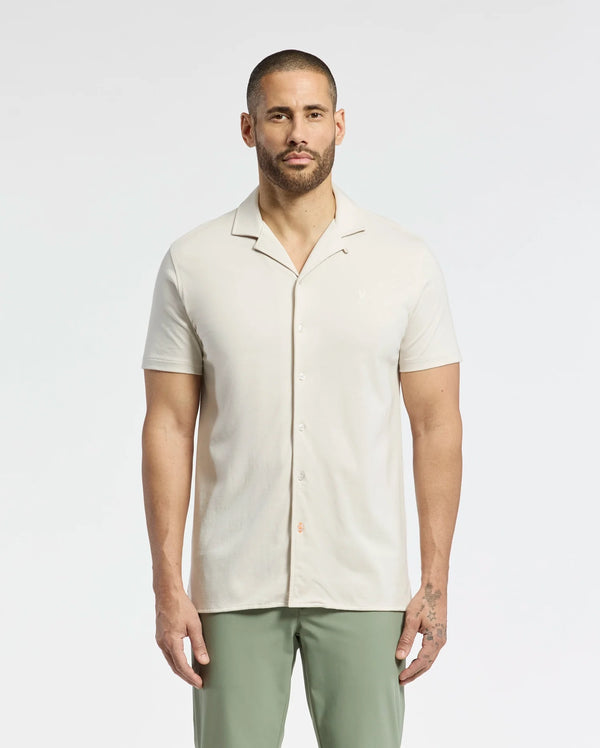 A person stands wearing a Psycho Bunny MENS BRYAN CAMP COLLAR SHORT SLEEVE SHIRT, crafted from a Pima Cotton Blend, with green pants. The plain white background highlights their casual look enhanced by the comfort of moisture-wicking performance fabric.
