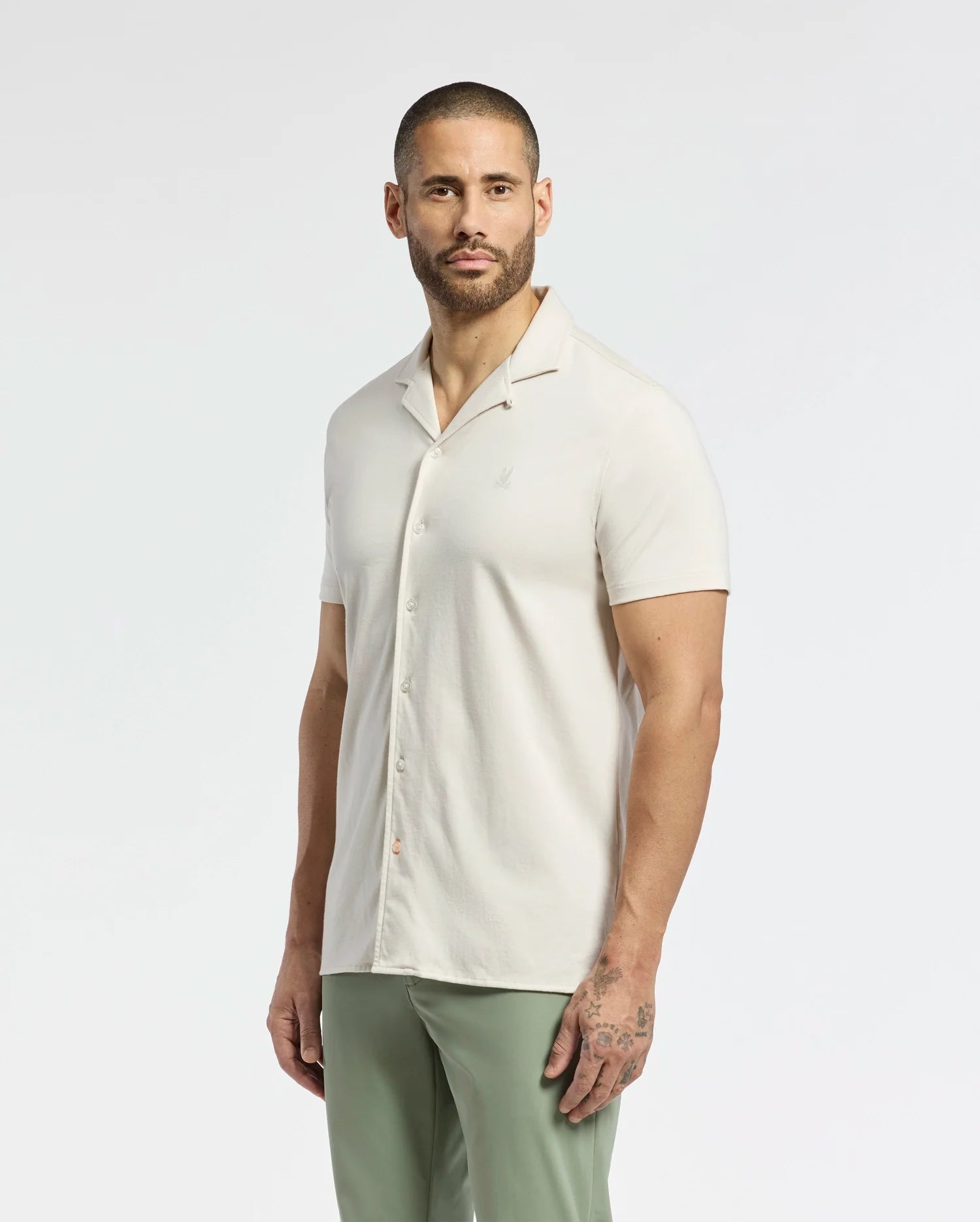 A person with a beard and short hair tilts their head slightly while wearing Psycho Bunny's Mens Bryan Camp Collar Short Sleeve Shirt (B6Q581F200) paired with light green pants, standing against a plain white background.