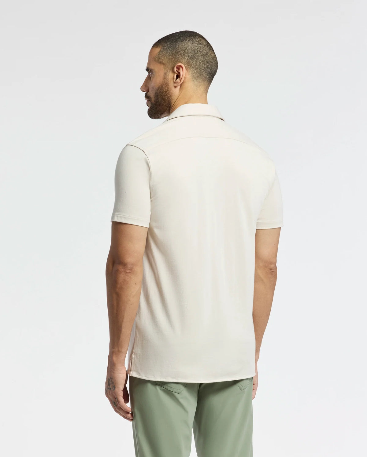 A person with short hair and a beard, facing away, wears a light beige MENS BRYAN CAMP COLLAR SHORT SLEEVE SHIRT by Psycho Bunny, made from moisture-wicking fabric, paired with green pants. A plain white background highlights the simple yet stylish outfit.