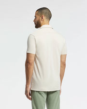 A person with short hair and a beard, facing away, wears a light beige MENS BRYAN CAMP COLLAR SHORT SLEEVE SHIRT by Psycho Bunny, made from moisture-wicking fabric, paired with green pants. A plain white background highlights the simple yet stylish outfit.