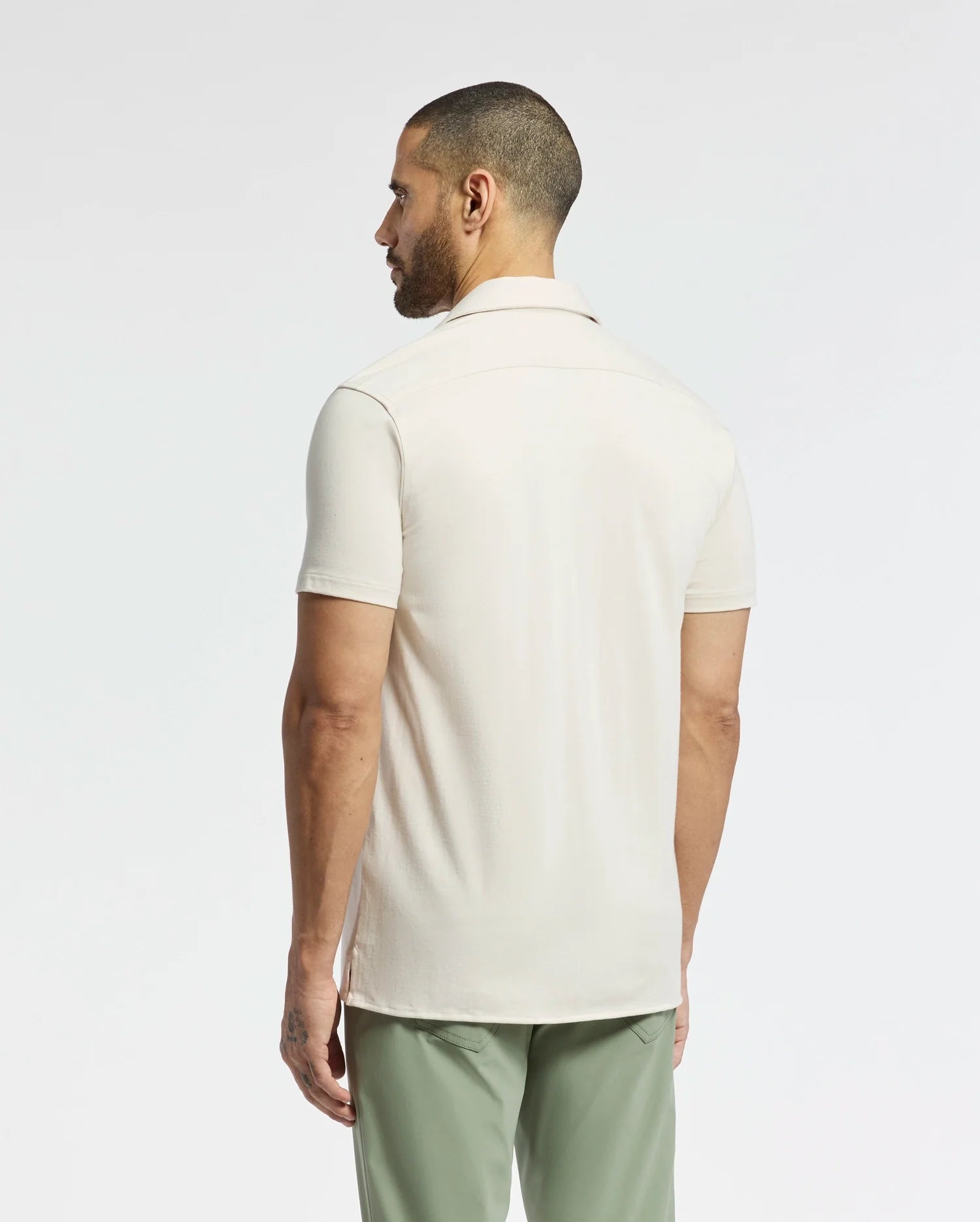 A person with short hair and a beard, facing away, wears a light beige MENS BRYAN CAMP COLLAR SHORT SLEEVE SHIRT by Psycho Bunny, made from moisture-wicking fabric, paired with green pants. A plain white background highlights the simple yet stylish outfit.