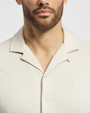 A man wears the Psycho Bunny MENS BRYAN CAMP COLLAR SHORT SLEEVE SHIRT in light beige, crafted from a Pima cotton blend, with an embroidered chest logo. The photo highlights the collar and upper chest against a clean background.