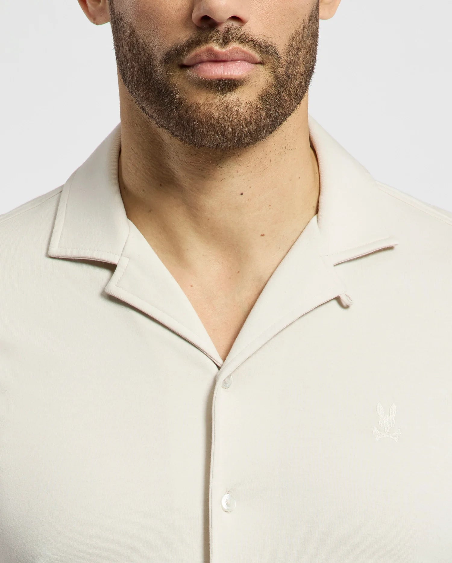 A man wears the Psycho Bunny MENS BRYAN CAMP COLLAR SHORT SLEEVE SHIRT in light beige, crafted from a Pima cotton blend, with an embroidered chest logo. The photo highlights the collar and upper chest against a clean background.