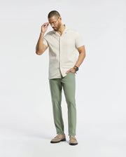 A man in a Psycho Bunny MENS BRYAN CAMP COLLAR SHORT SLEEVE SHIRT (B6Q581F200), crafted from moisture-wicking Pima cotton blend, stands against a white background. He adjusts his glasses with one hand while wearing green pants, beige loafers, and a watch.