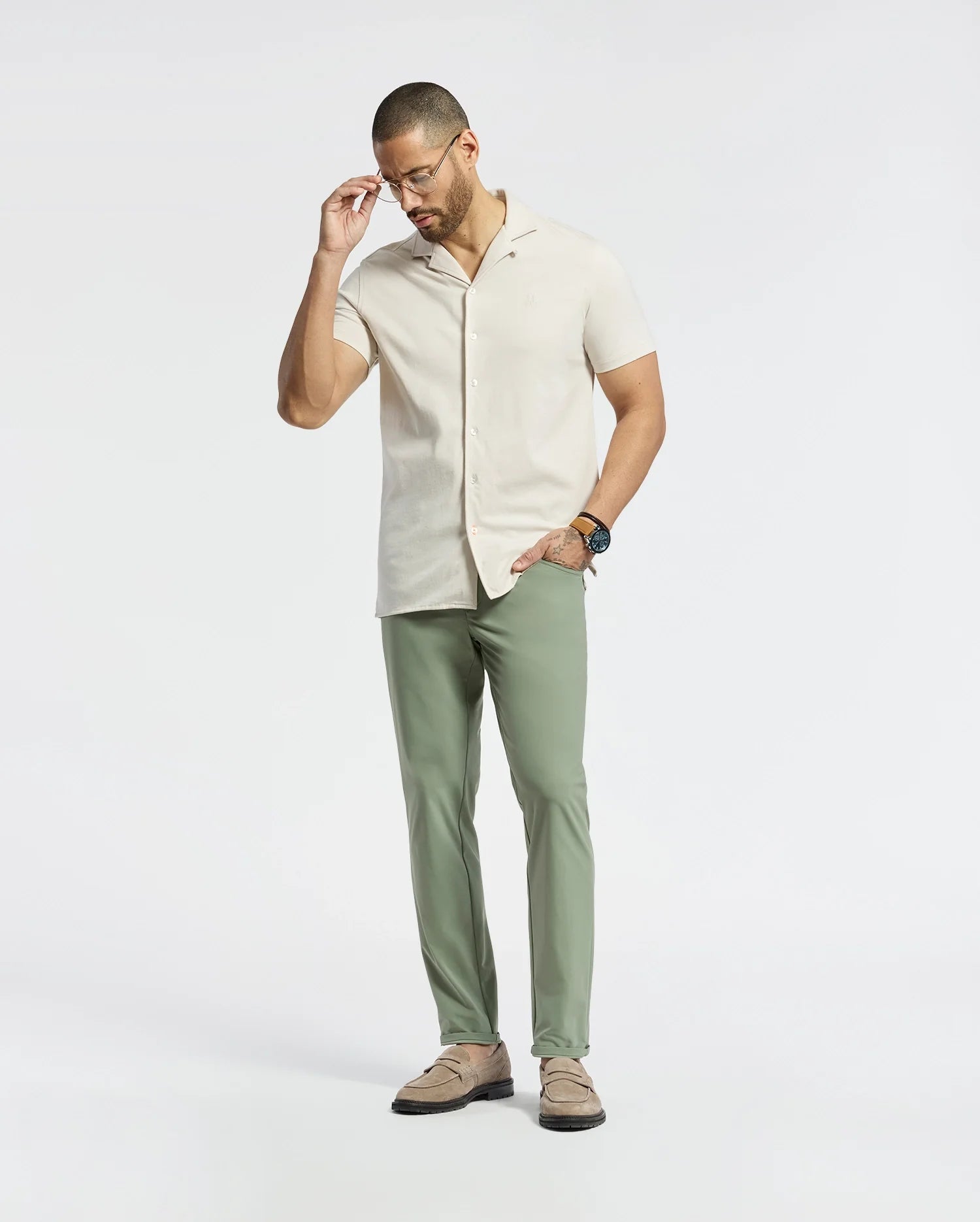 A man in a Psycho Bunny MENS BRYAN CAMP COLLAR SHORT SLEEVE SHIRT (B6Q581F200), crafted from moisture-wicking Pima cotton blend, stands against a white background. He adjusts his glasses with one hand while wearing green pants, beige loafers, and a watch.