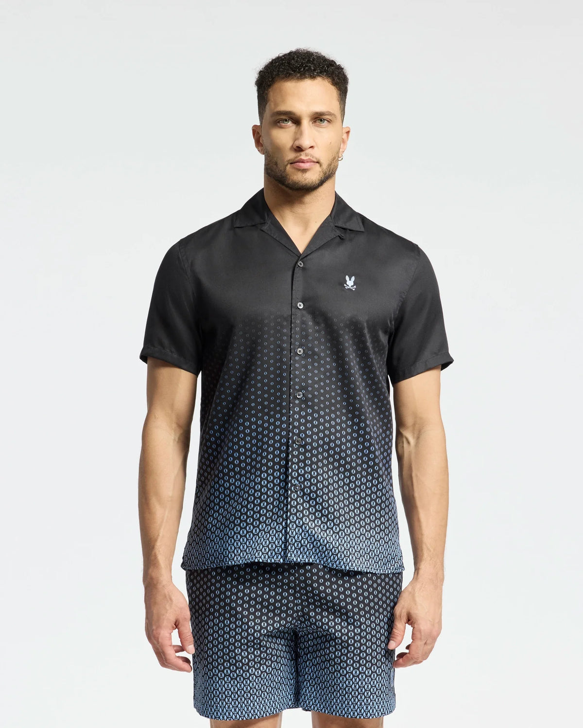 A man models Psycho Bunny's MENS BENDIGO TENCEL SHIRT, a black short-sleeve button-up with matching shorts. Its subtle ombre effect and white dots that thicken toward the bottom enhance its style, while the camp collar provides a casual contrast against a plain, light background.