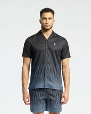 A man models Psycho Bunny's MENS BENDIGO TENCEL SHIRT, a black short-sleeve button-up with matching shorts. Its subtle ombre effect and white dots that thicken toward the bottom enhance its style, while the camp collar provides a casual contrast against a plain, light background.