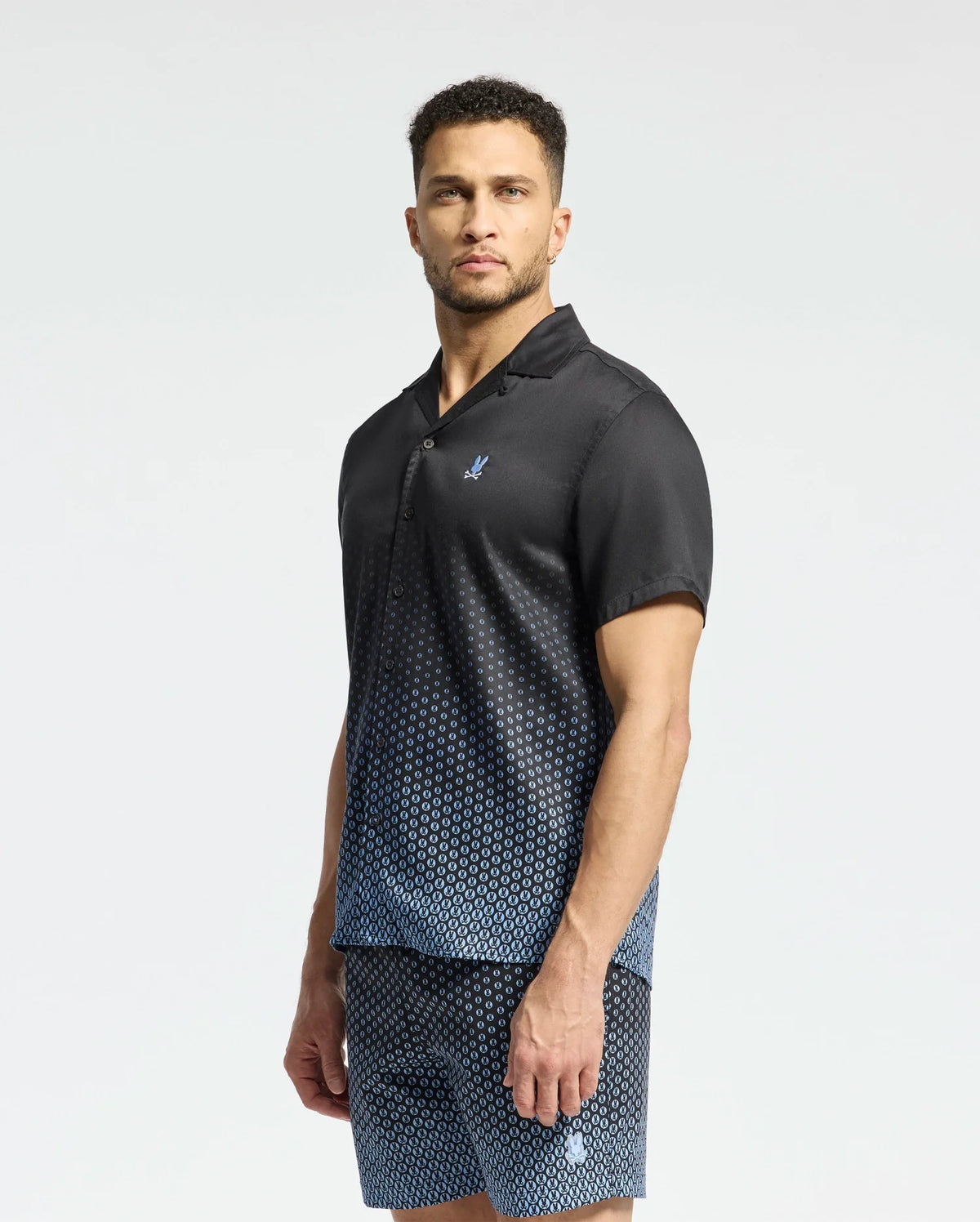 A man stands with a neutral expression against a plain background, wearing a dark Psycho Bunny MENS BENDIGO TENCEL SHIRT - B6Q627F200 featuring an ombre effect and small chest logo, paired with matching shorts.