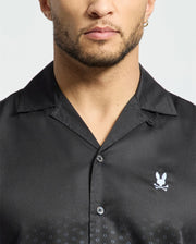 Donning the Psycho Bunny MENS BENDIGO TENCEL SHIRT - B6Q627F200, featuring a sleek black design with a camp collar and iconic white rabbit and crossed bones logo, complements their facial hair and earring, adding an edgy touch to their style.