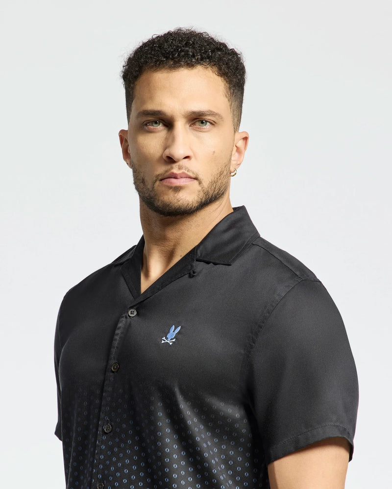 A person with short curly hair wears the MENS BENDIGO TENCEL SHIRT by Psycho Bunny, a black short-sleeved shirt featuring a small blue logo on the chest and a subtle ombre effect. The background is plain white.