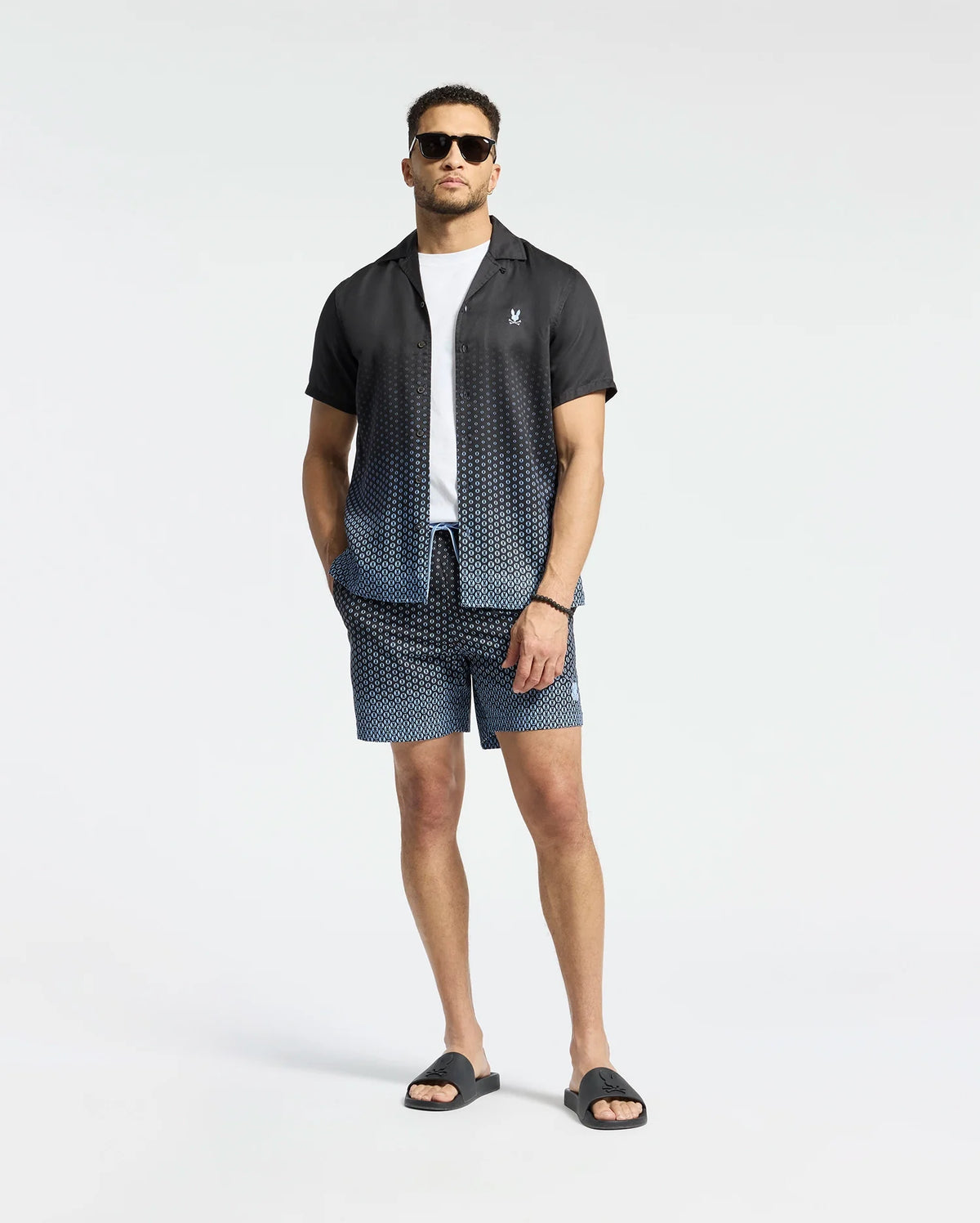 A man in Psycho Bunny's MENS BENDIGO TENCEL SHIRT, featuring a black ombre design and camp collar, stands confidently in matching shorts. A white T-shirt underneath and black slide sandals polish off his look against a plain white backdrop.