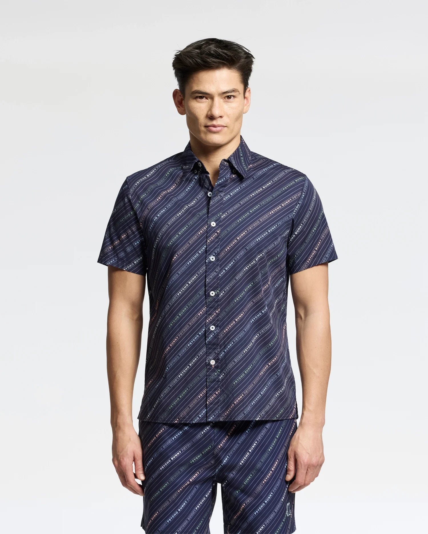 A person wearing the Psycho Bunny MENS RENMARK stretch poplin woven shirt in dark blue with a logo-striped pattern stands against a plain background.